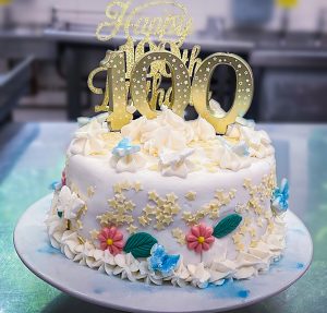 Celebratory 100th Birthday Cake. The cake is white and is decorated with pink and blue iced flowers. On top of the cake are numbered candles that spell '100'