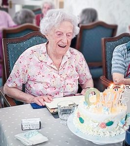 Image of 100 year old, Joyce Douglas, enjoying her 100th birthday at Redcot Residential Care Home. In front of Joyce is a white birthday with iced decorations and lit birthday candles
