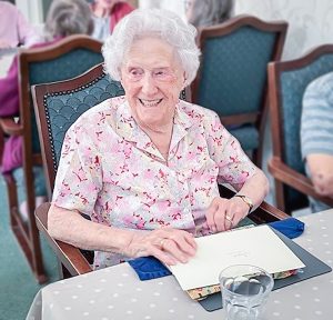 Image of Joyce Douglas at Redcot residential care home. Joyce is wearing a pink and white blouse and is smiling.